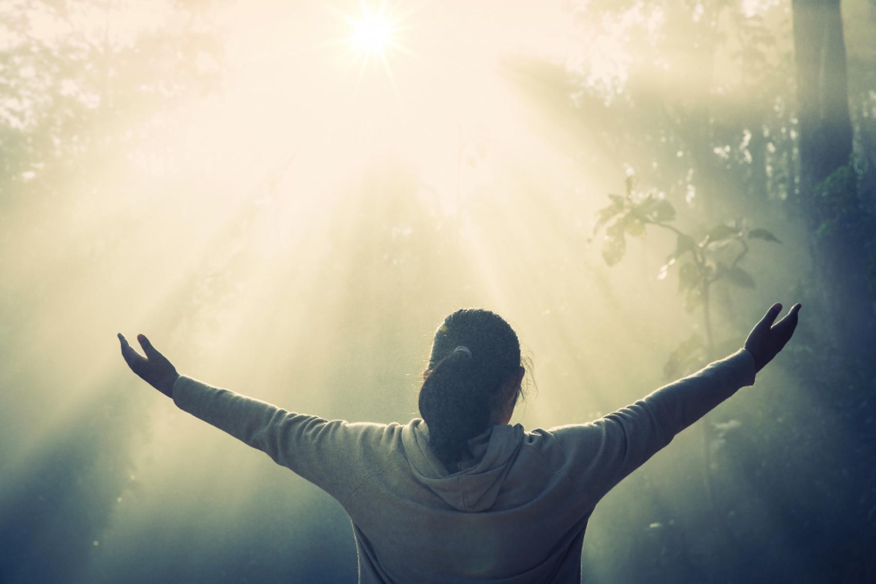 Sunlight shining into a woodland. Person with long hair in a ponytail facing the light and lifting their arms up towards it.