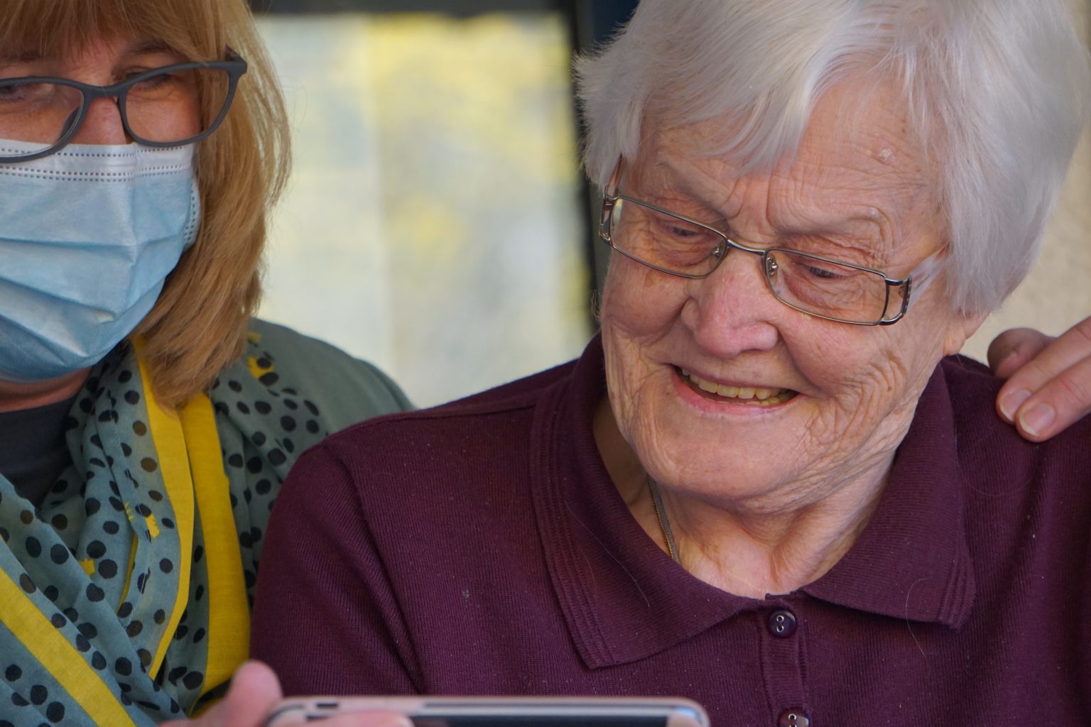Care worker in a face mask sitting beside a smiling older woman looking at a phone