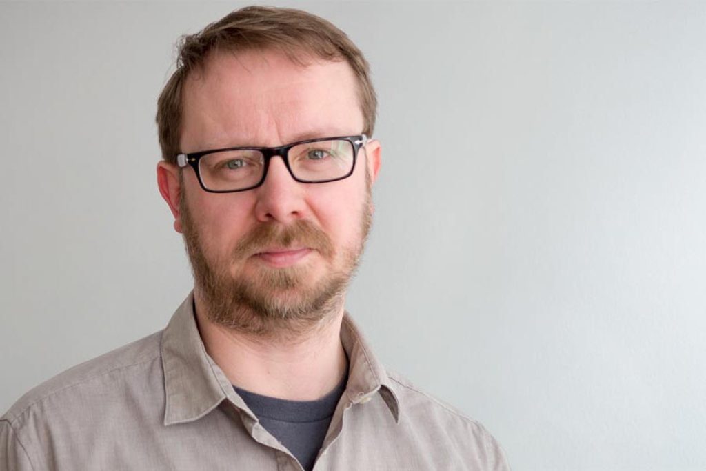 Ian Phillip with glasses and a beard, in a grey shirt